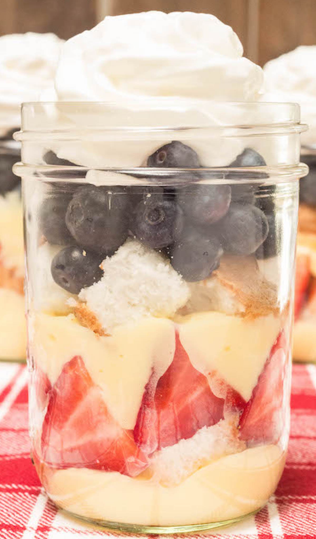 A small mason jar filled with layers of pudding, strawberries, angel food cake, blueberries, and whipped cream on a red picnic style table cloth.