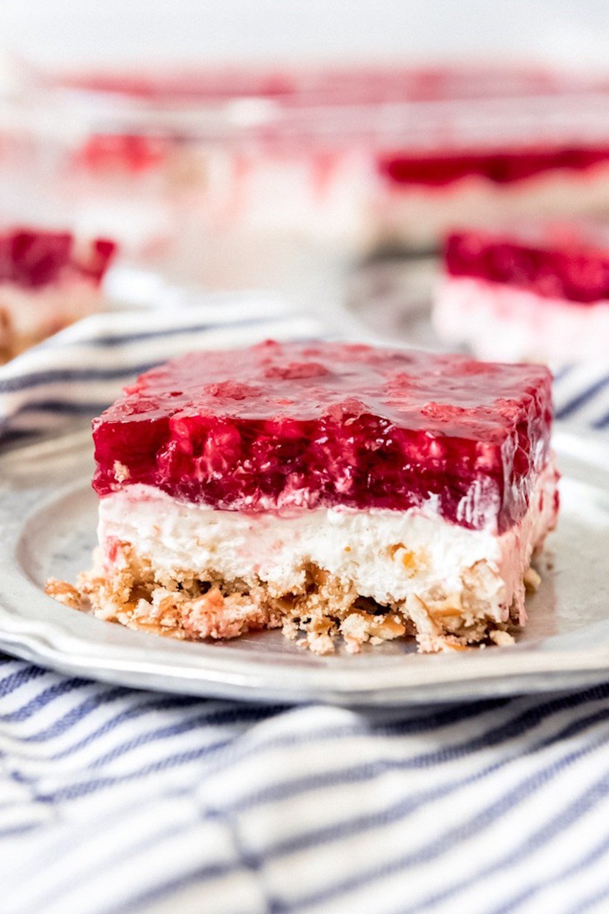 A cut square of raspberry pretzel salad sits on a serving plate with the rest of the pretzel salad out of focus in the background.