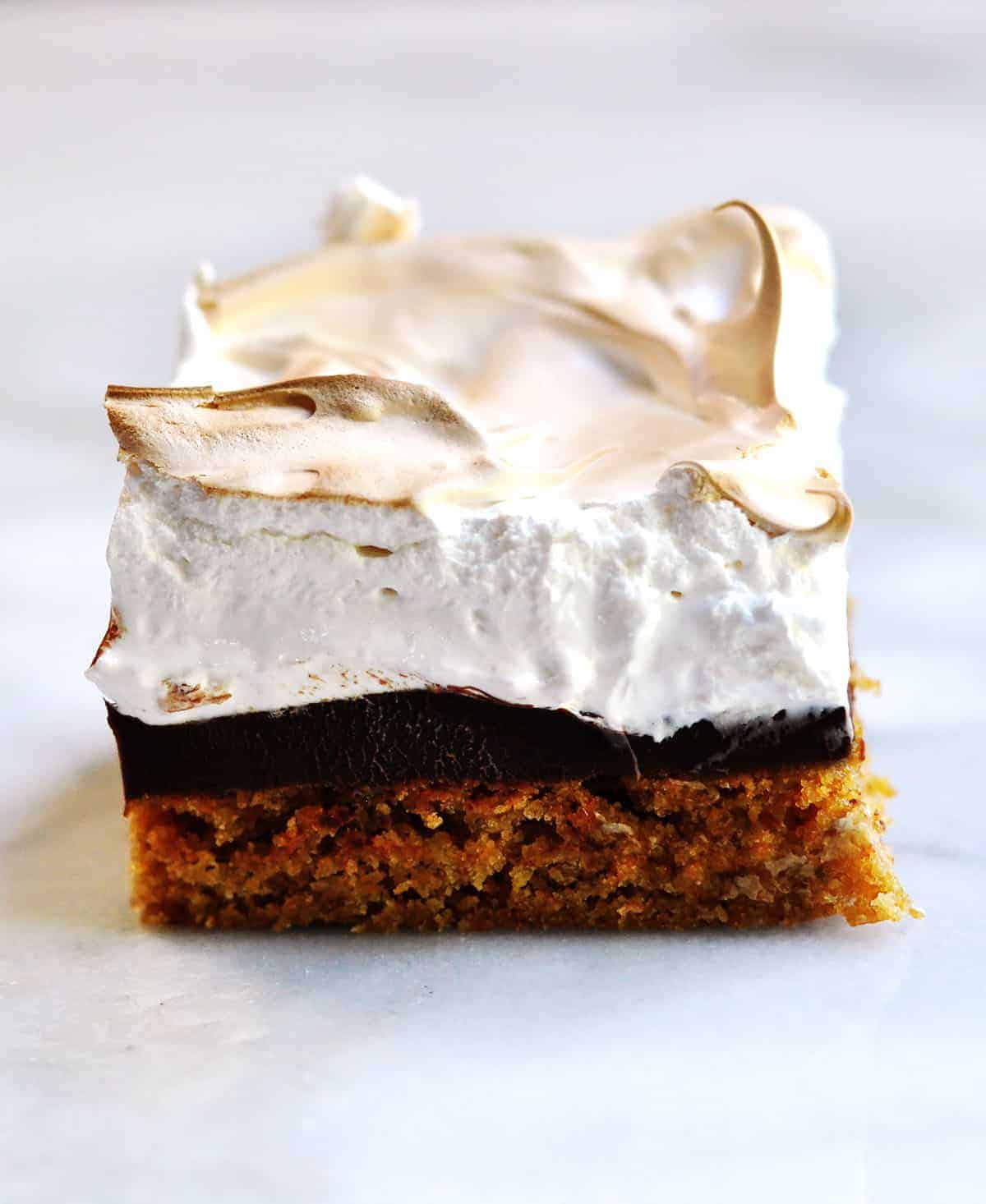 A brownie with a clear graham cracker layer, chocolate layer, and toasted marshmallow meringue layer sits on a white background.