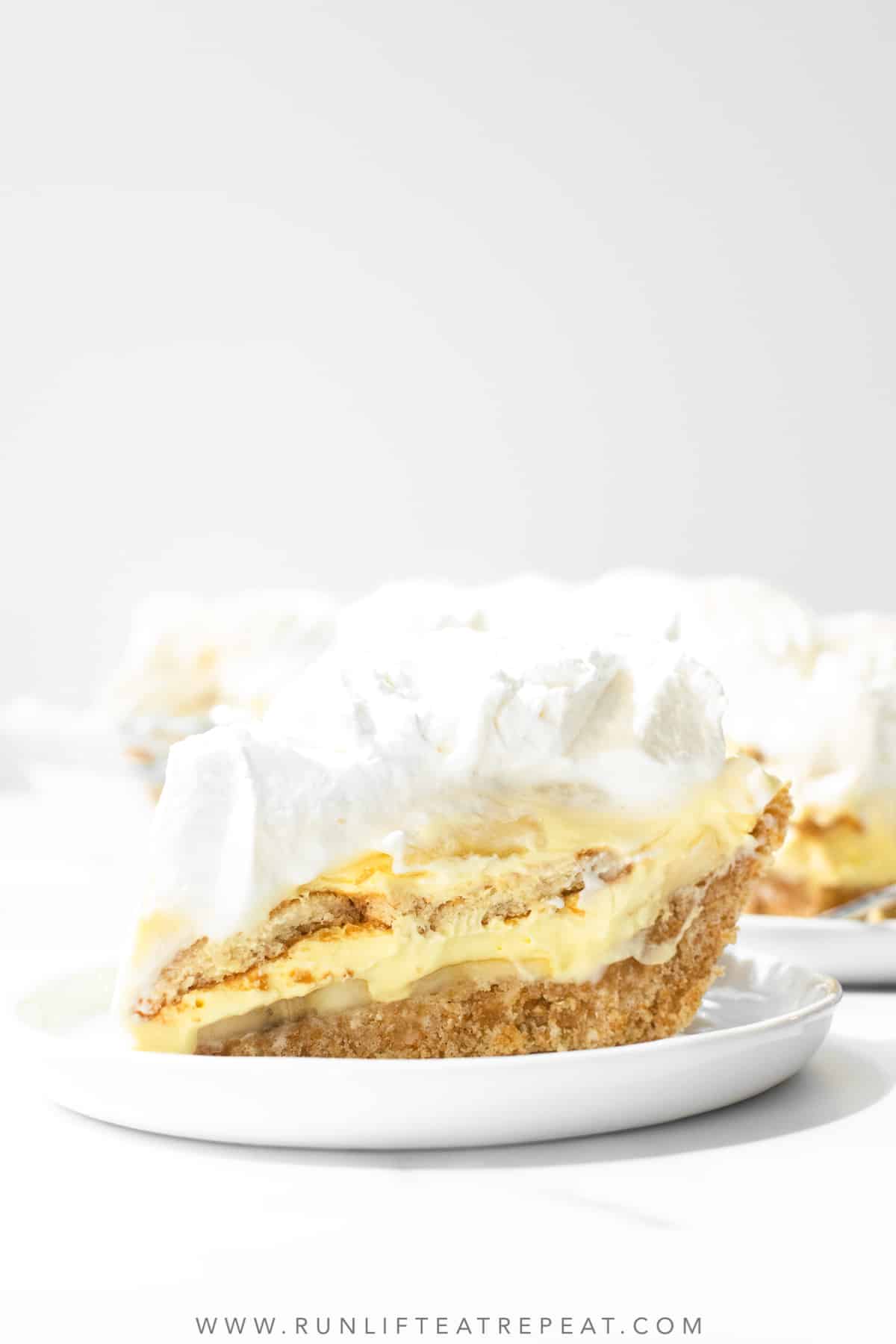 A single slice of banana pudding pie sits on a white serving plate in front of the rest of the pie.