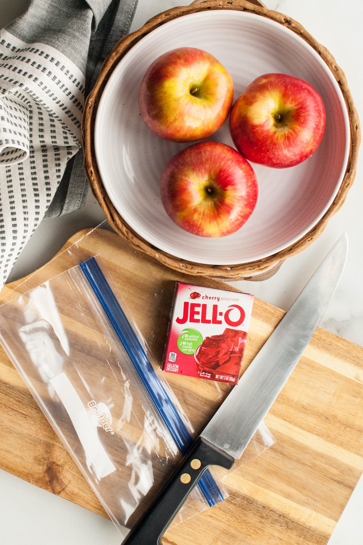 Overhead view of jello apple ingredients (3 apples, 1 box of jello powder) sitting next to a knife, ziploc bag, and a cutting board.