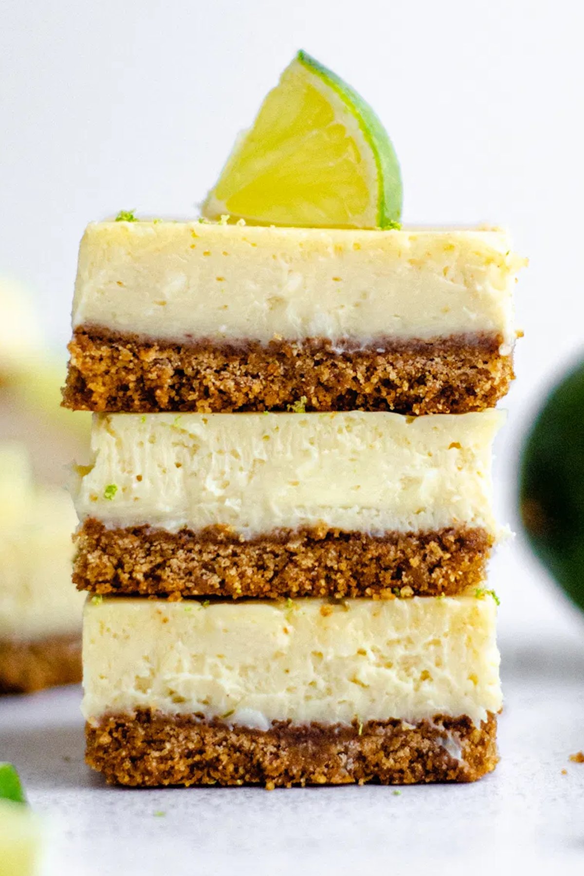 Three key lime pie bars stacked on top of one another on a light background. The top bar is garnished with a lime slice.