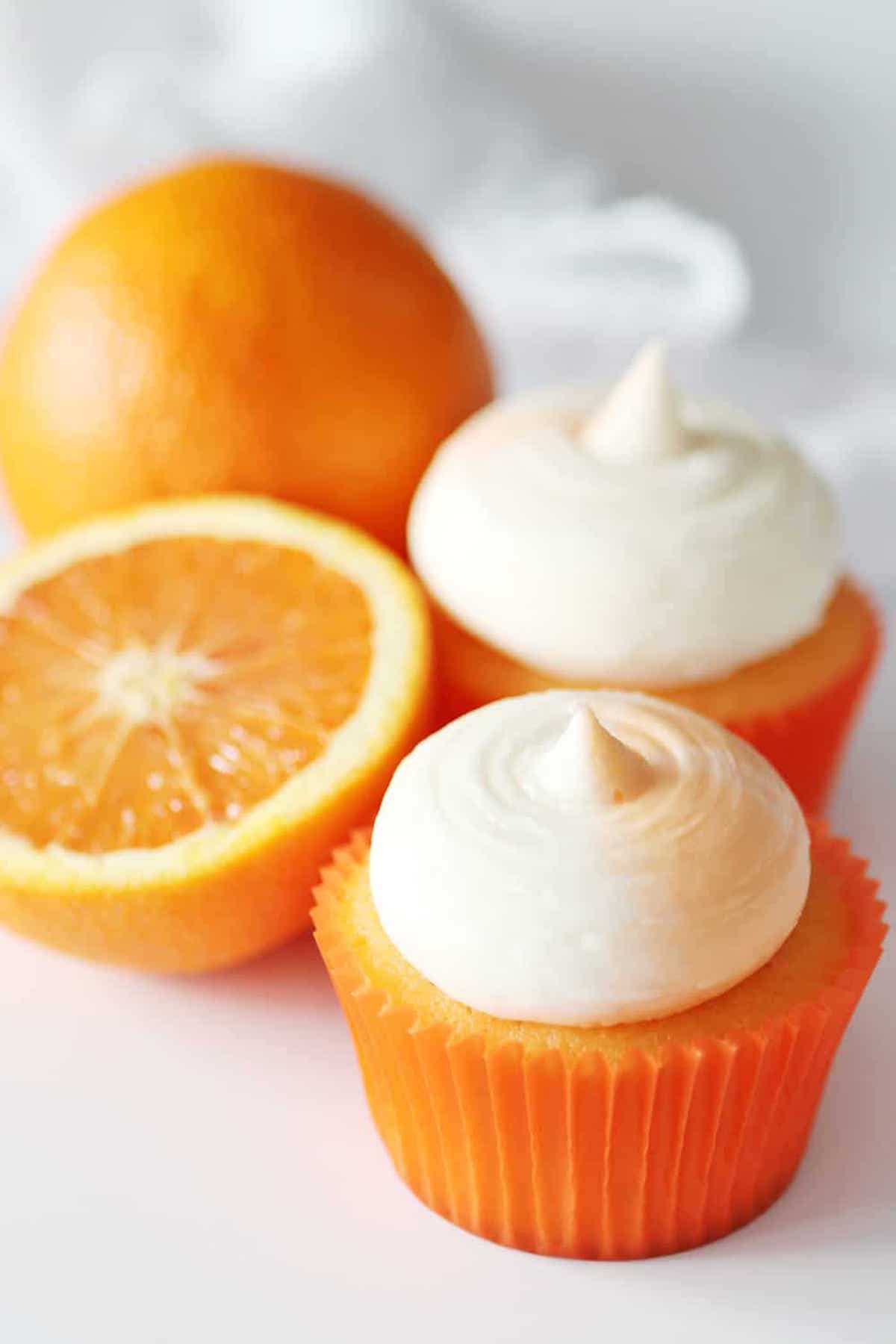 Two orange creamsicle cupcakes topped with white frosting sit on a light background next to an orange that has been sliced in half.