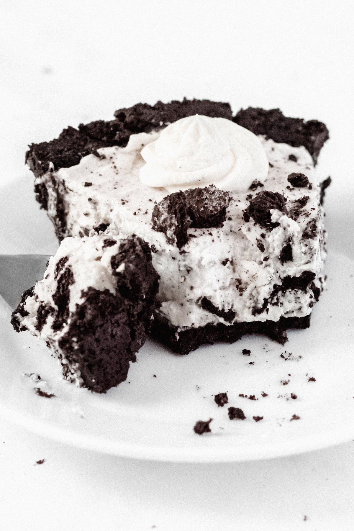 A slice of cookies & cream pie with a forkful taken out of it sits on a white plate on a white background