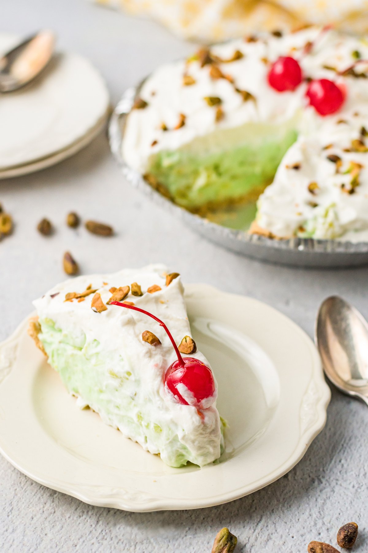 A slice of pistachio pie garnished with a maraschino cherry sits on a small serving plate in front of the rest of the pie.