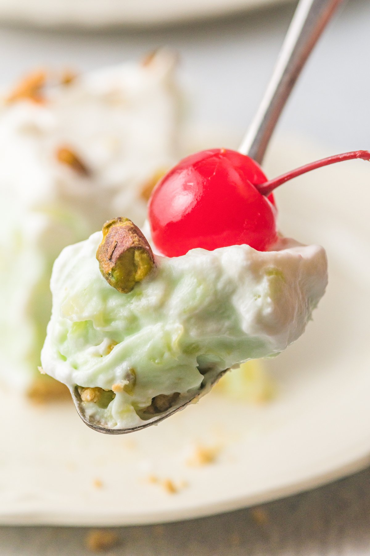A spoon holds a bite of the pistachio pie and a maraschino cherry. The piece of pie itself is blurred out in the background.