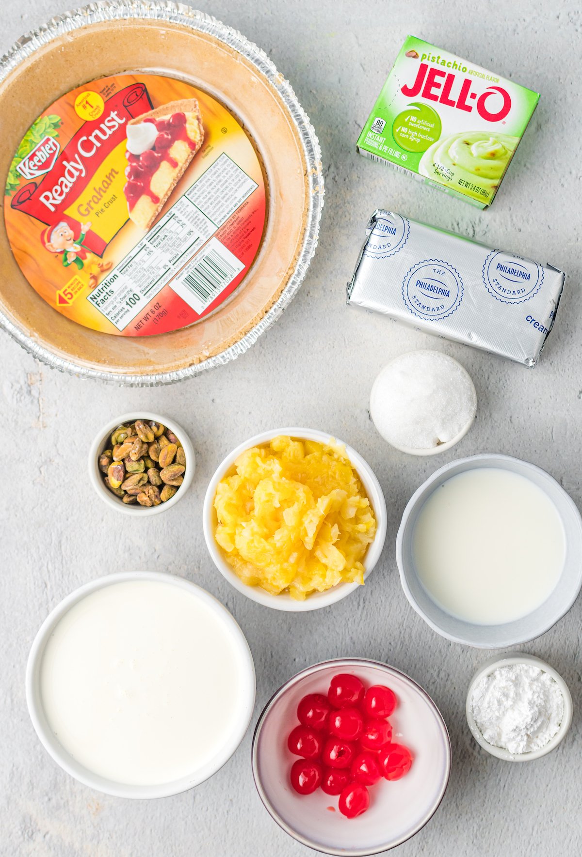 Overhead view of pie ingredients: store-bought graham cracker crust, Jello Pistachio pudding mix, cream cheese, crushed pineapple, sugar, maraschino cherries, chopped pistachios, milk, and heavy cream.