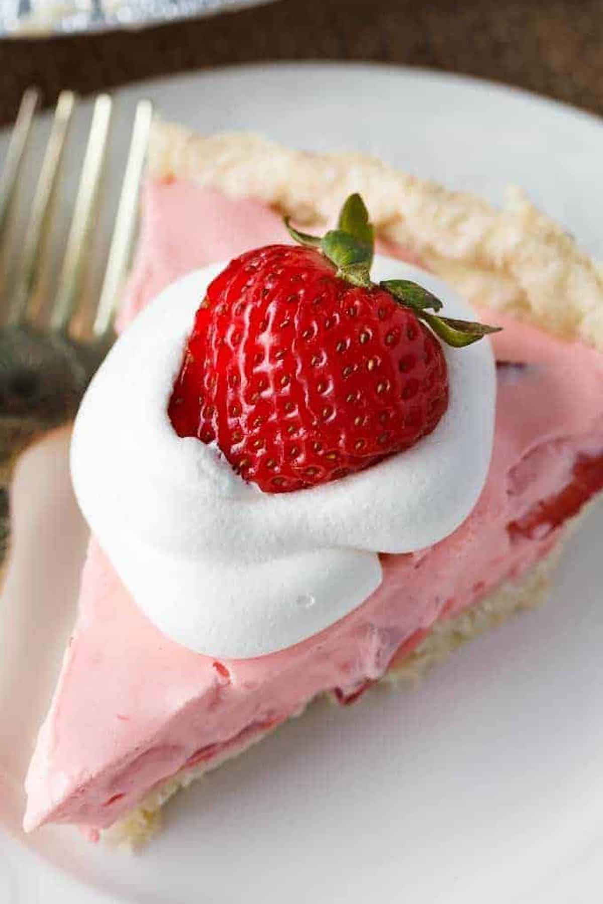 A slice of pink strawberry cream pie garnished with whipped cream and a whole strawberry sits on a white plate.