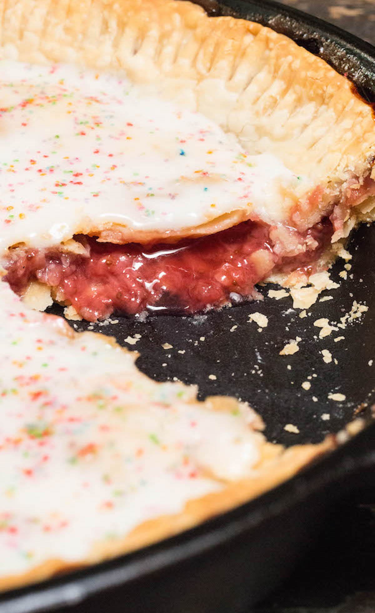 A cast iron skillet is filled with a strawberry pie that is glazed like a pop-tart. One slices has been removed to show the strawberry filling inside.