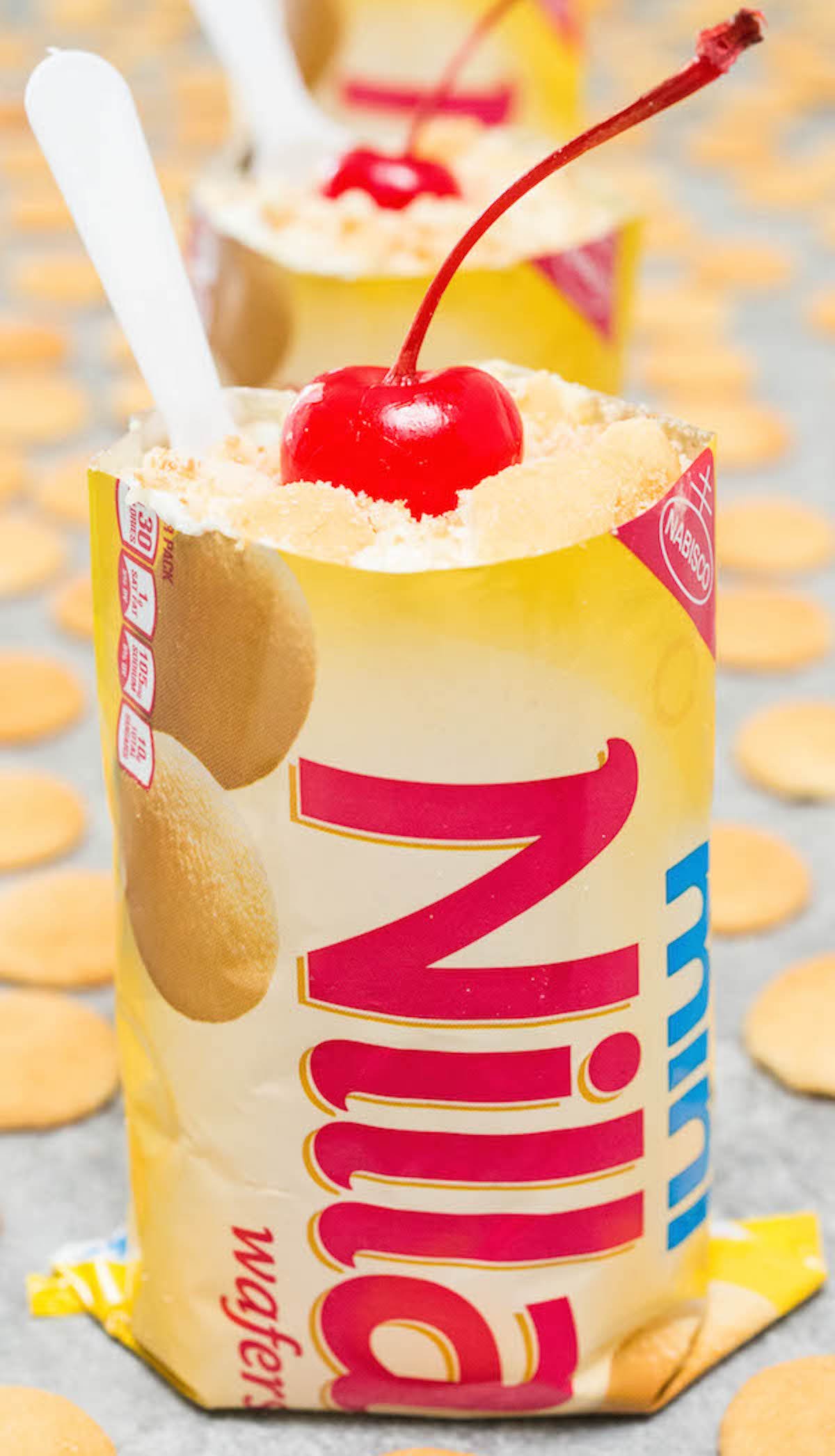 A small bag of mini nilla wafers is filled with banana pudding and topped with a maraschino cherry and small plastic spoon.
