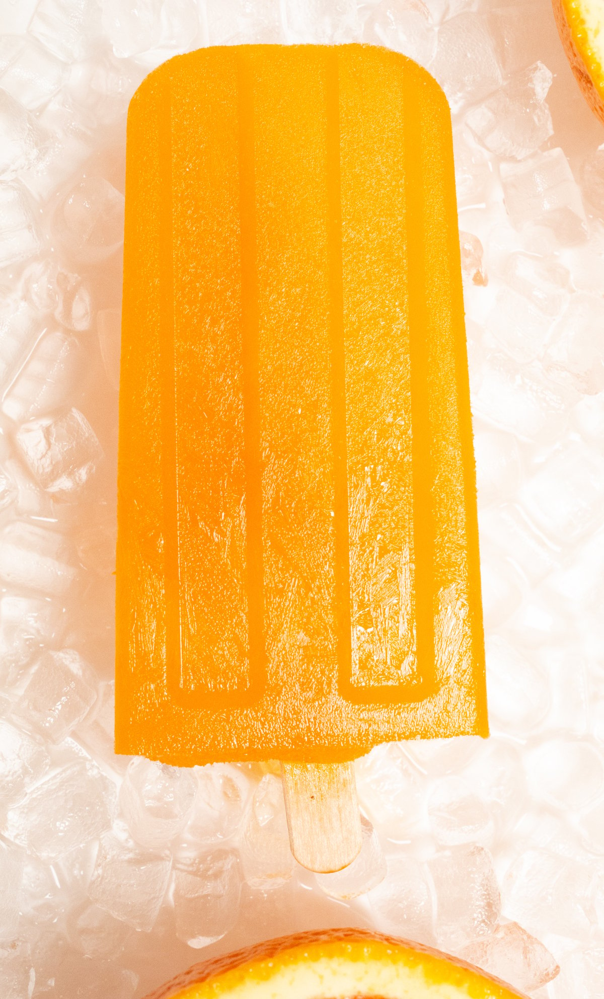 A single orange popsicles lays on a bed of pebble ice on a white background.