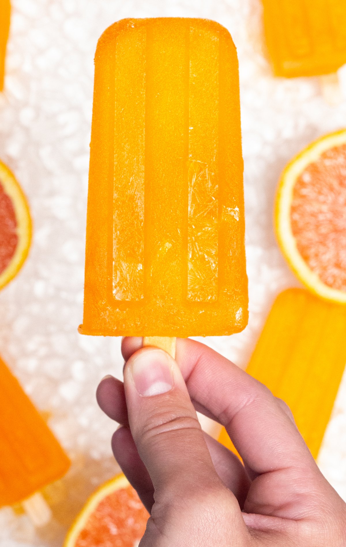 A hand holds up an Orange Popsicle in front of a white background. A few other orange popsicles are in the background out of focus.