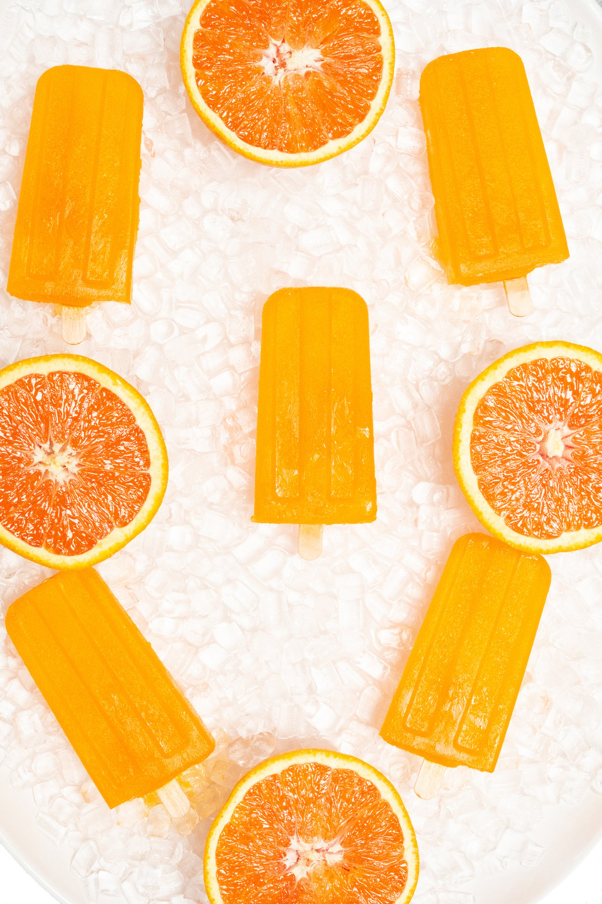 Five orange popsicles are arranged on pebble ice on a white platter. They are garnished with oranges sliced in half.