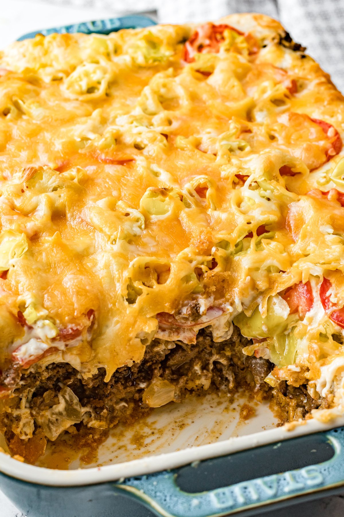 A blue Staub casserole dish filled with John Wayne casserole has a piece cut out to show the ground beef, vegetable, and cheese layers.