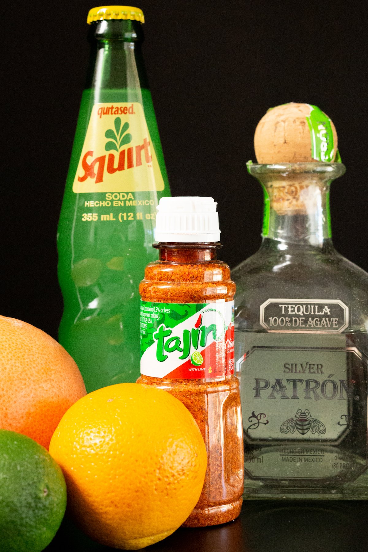 All the ingredients to make a cantarito on a black background: a fresh grapefruit, an orange, a lime, tajin chile salt, squirt soda in a glass bottle, and a bottle of patron.