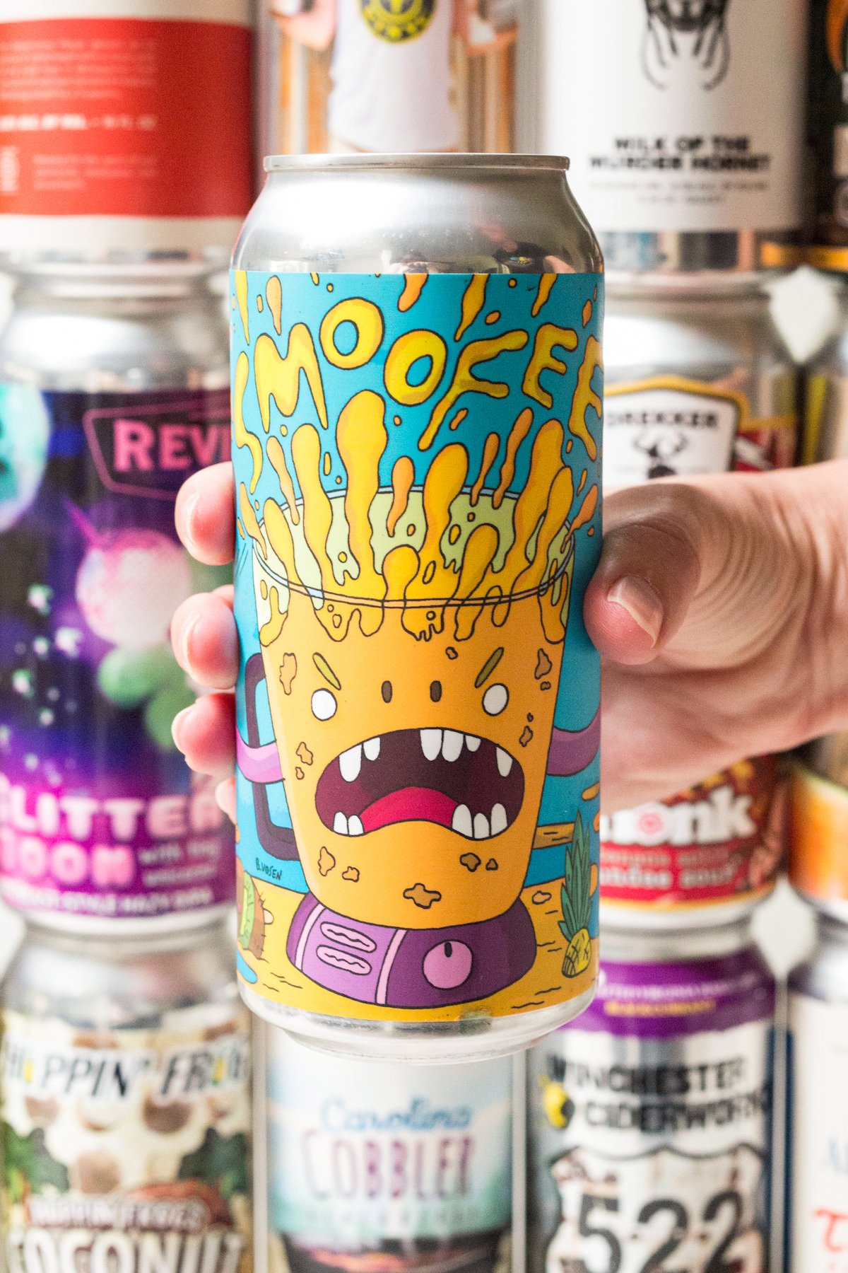 A hand holds a "Smoofee" beer in front of a wall of beers. The smoofee beer's label features a blender with eyes and a mouth full of orange liquid.