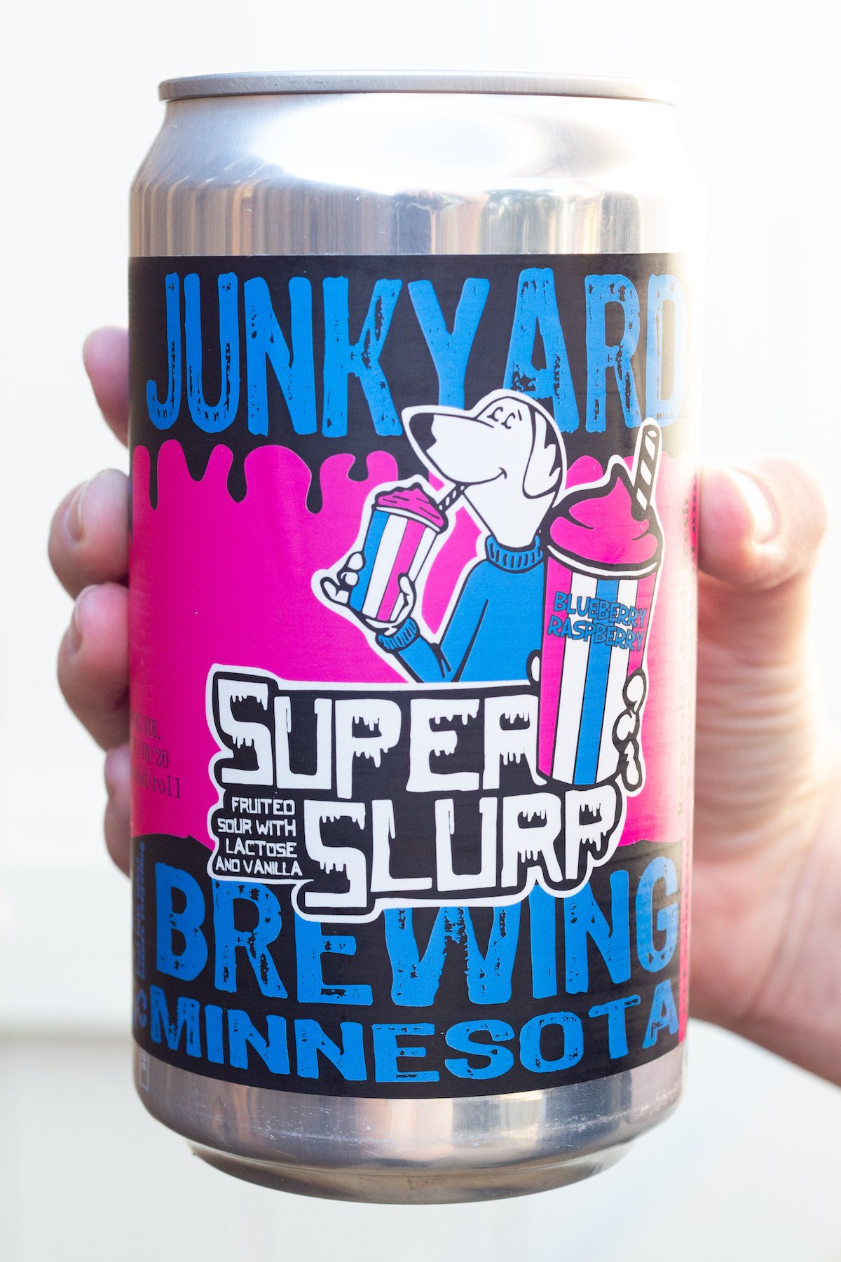 A hand holds up a can of Junkyard Super Slurp sour beer against a white background. The label of the beer is pink and blue and has a polar beer mascot holding a slurpee.