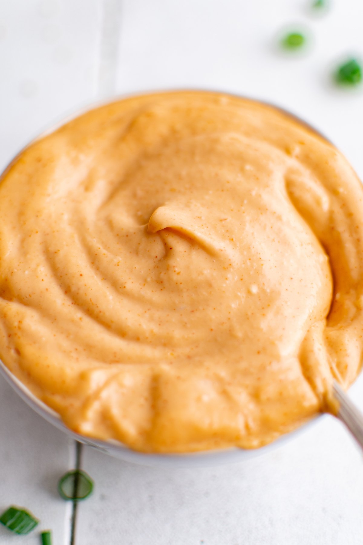 A white bowl filled with a slightly orange "yum yum sauce" that sits on a white tile background.