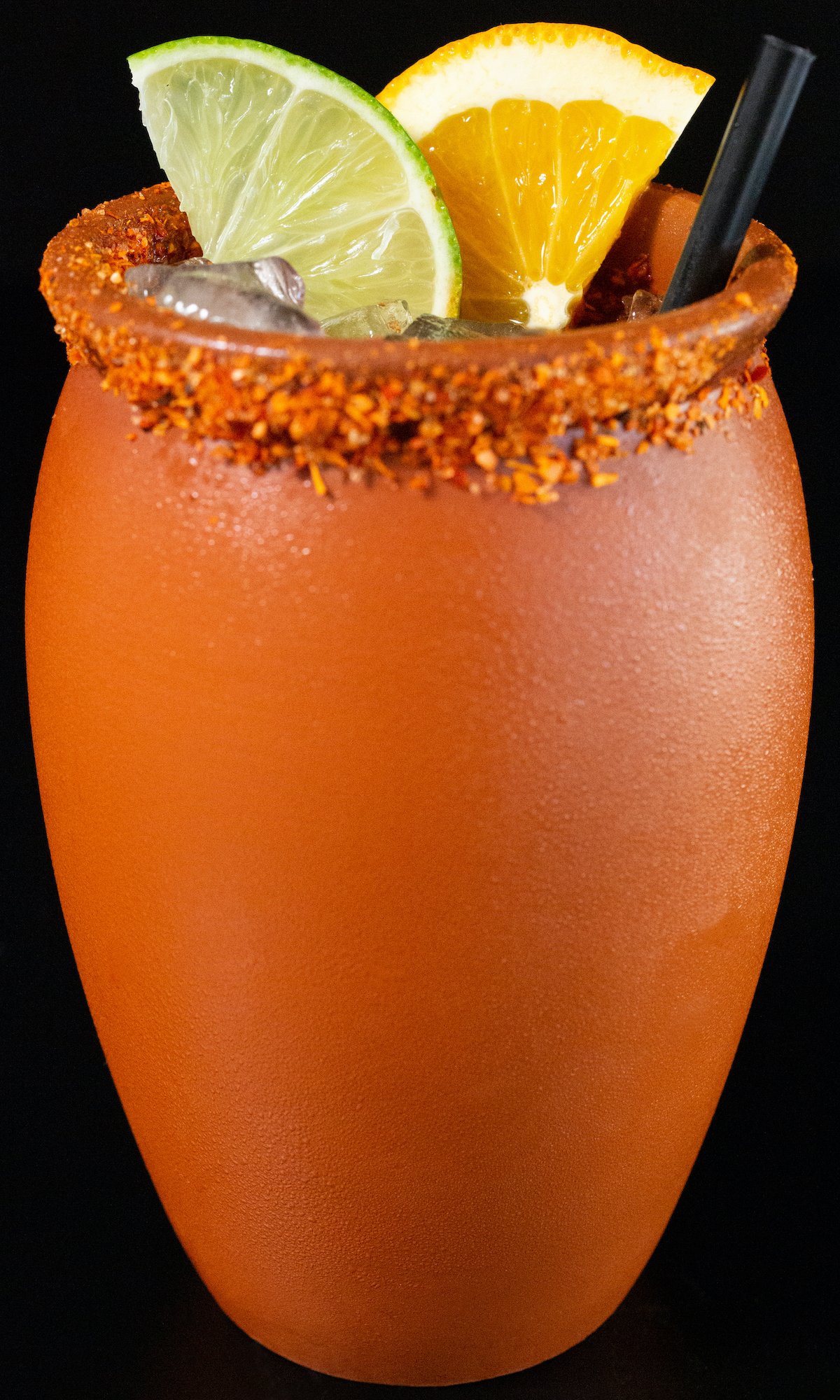 A clay pot filled with a cantarito cocktail sits on a black background. The rim of the pot is covered in chile salt and it is garnished with a lime and orange slice.