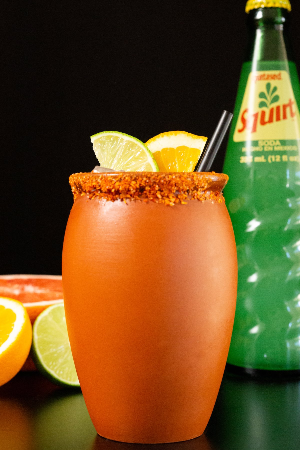 A clay jar that's filled with a cantarito cocktail is garnished with a lime & orange slice. It sits on a black background next to a glass bottle of squirt soda and sliced grapefruit, orange, and lime.