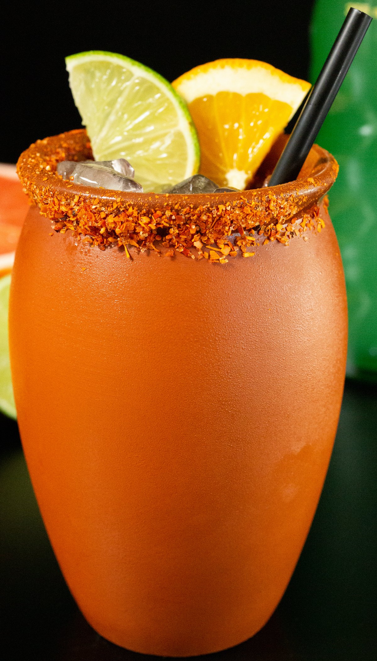 A clay glass with a chile salt rim, a lime slice, an orange slice, and a black straw sit on a black background.