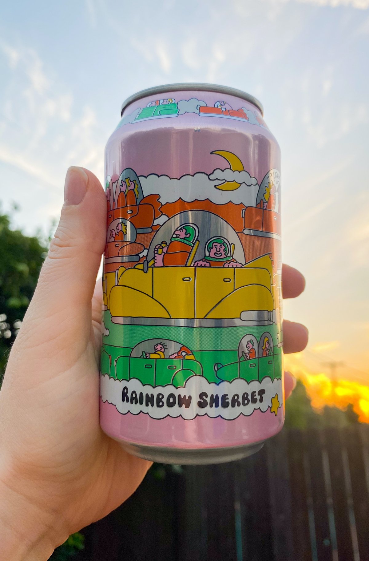 A hand holds a colorful can of rainbow sherbet beer against a sky and sunset background.