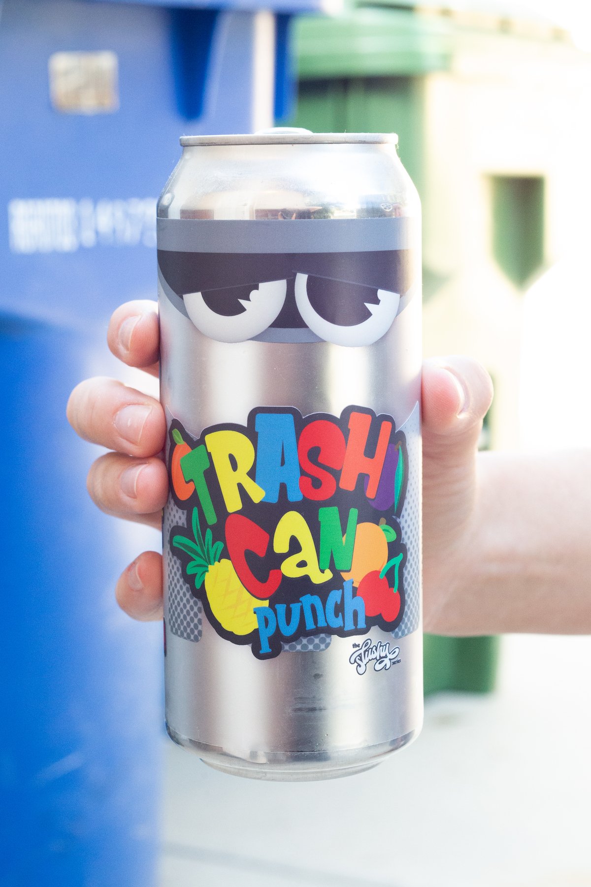 A hand holds a silver can of beer that reads "Trash Can Punch" in front of trash cans. 