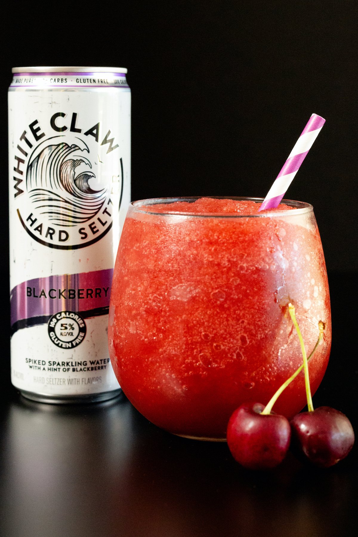 A stemless wine glass filled with a red white claw slushie sits in front of a White Claw can on a black background.