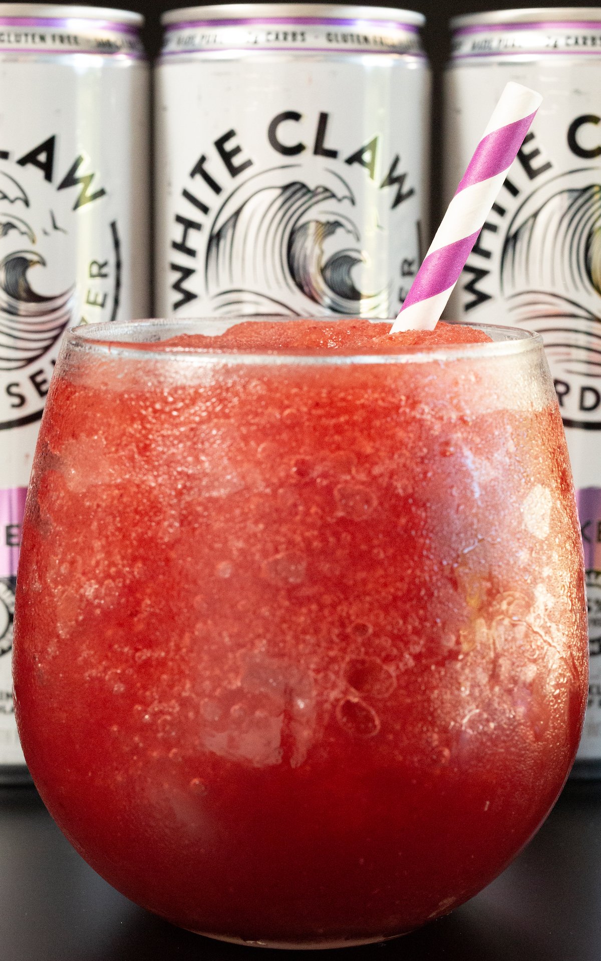 A close up of a stemless wine class filled with a red slushie in front of three cans of White Claw hard seltzer.