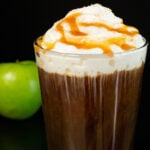 A pint glass filled with cold brew coffee and topped with whipped cream and caramel sits next to a green apple on a black background.