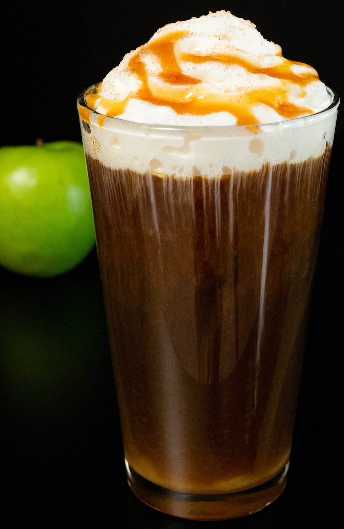 A pint glass filled with cold brew coffee and topped with whipped cream and caramel sits next to a green apple on a black background.