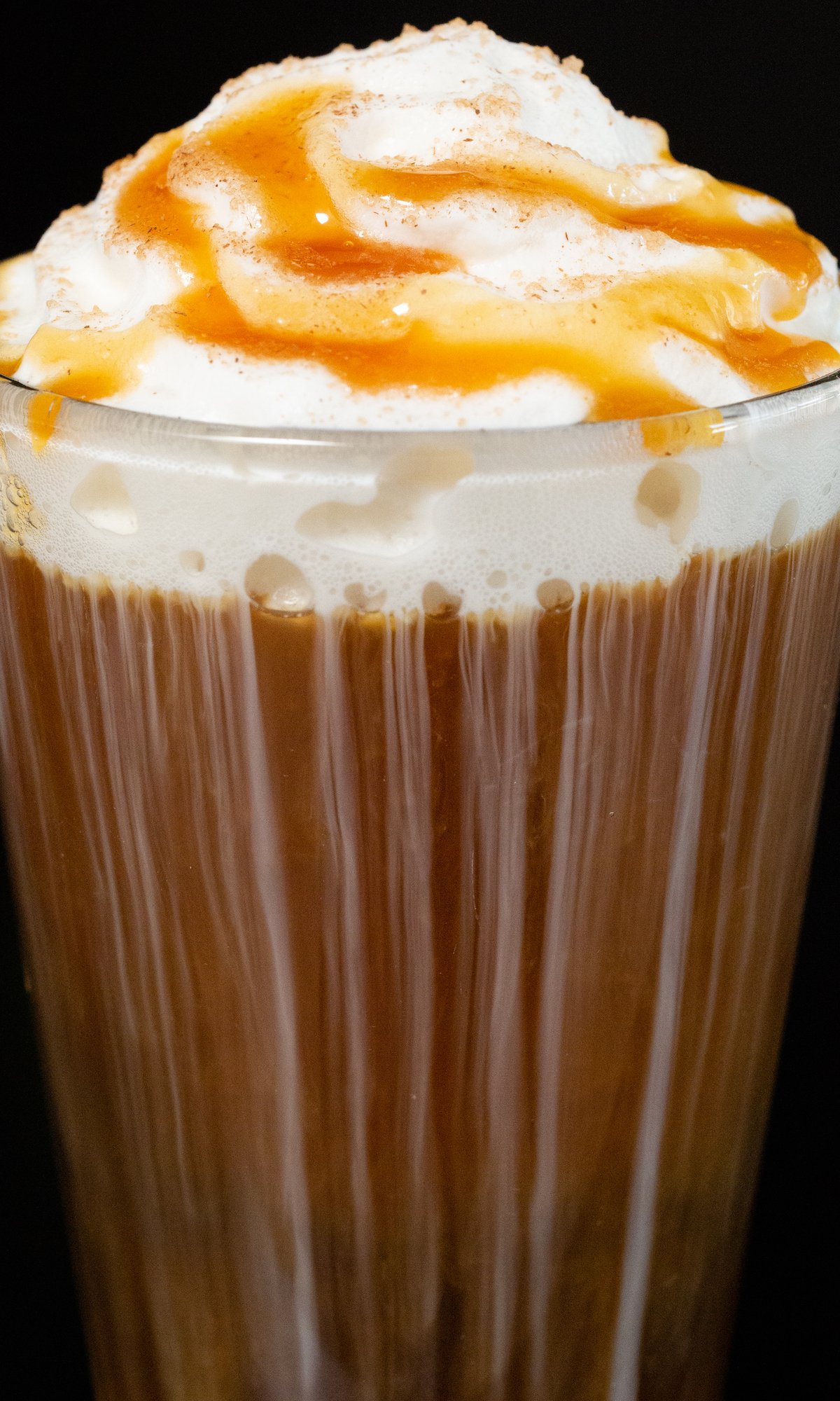 A close up of a caramel apple cold brew coffee topped with whipped cream and caramel sauce in a pint glass on a black background.