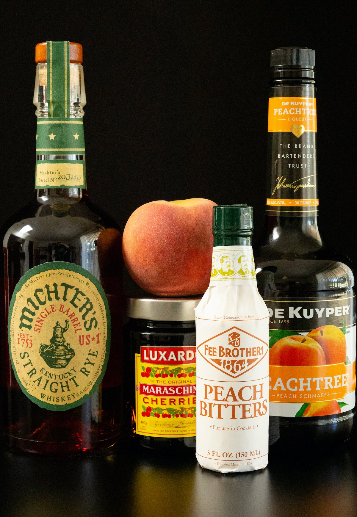 All the ingredients for peach old fashioned sit on a black background. A bottle of Michter's Rye Whiskey, maraschino cherries, peach schnapps, peach bitters, and a fresh peach.