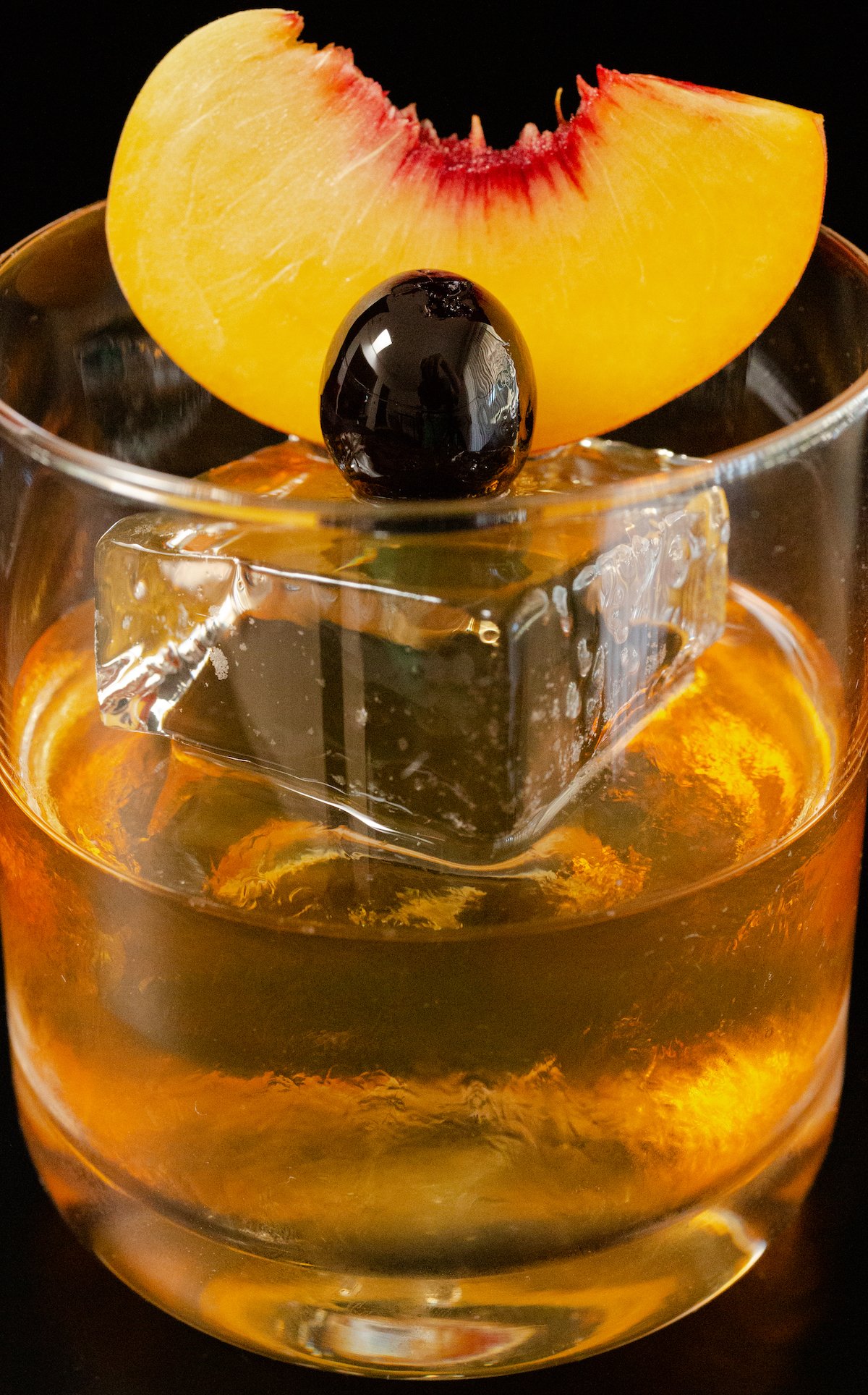 Close up of a clear lowball glass filled with a piece of large block ice, then filled halfway with old fashioned mixture. A peach slice and cherry are garnish.