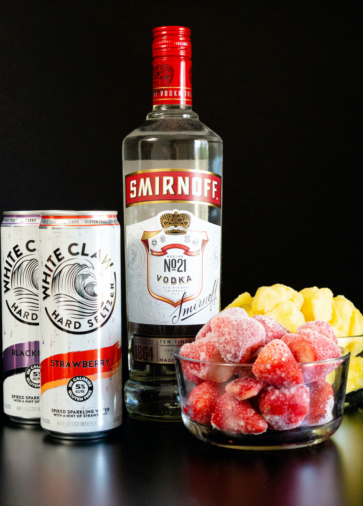 All the ingredients to make a white claw slushy sit on a black background. A bottle of smirnoff vodka, white claw cans, and frozen fruit.