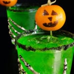 Two bright green Halloween margaritas with black salt rims and mandarin oranges with jack-o-lantern faces drawn on as garnishes sit on a black background.