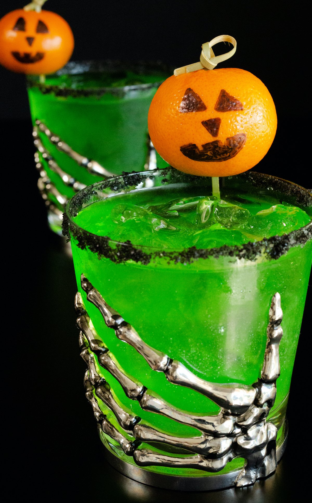 Two bright green Halloween margaritas with black salt rims and mandarin oranges with jack-o-lantern faces drawn on as garnishes sit on a black background.