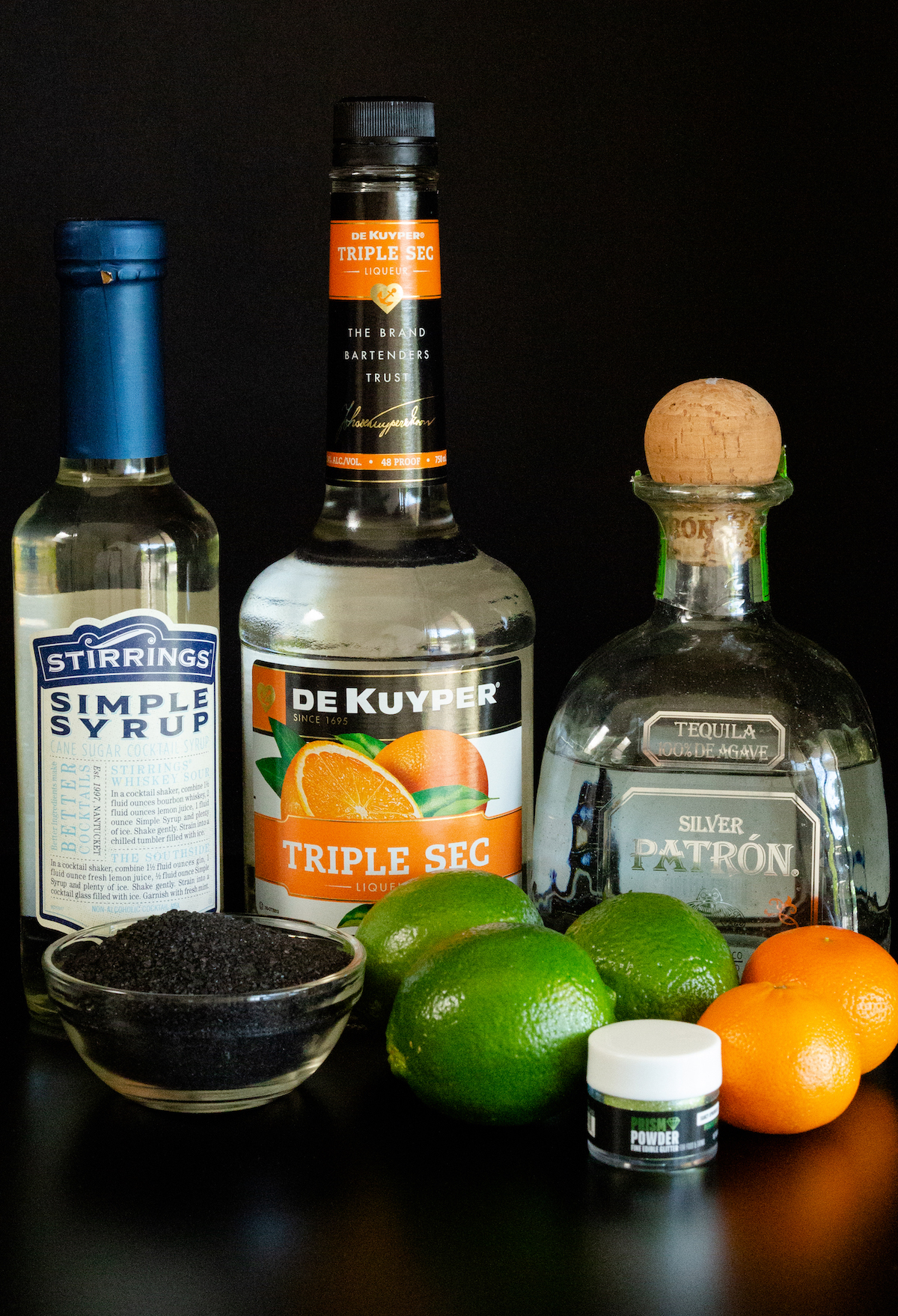 All the ingredients to make halloween margaritas sit on a black background. Simple syrup, triple sec, tequila, lava salt, limes, and small mandarin oranges.