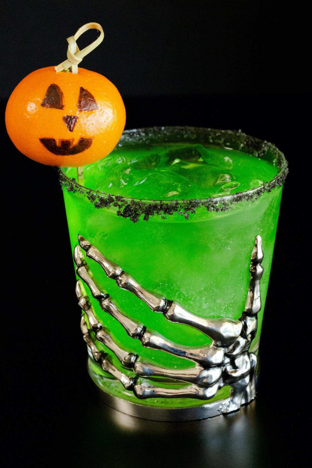 A lowball glass with a skeleton hand on the front is filled with a bright green margarita. There is a black salt rim and jack o lantern garnish.