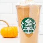 A tall plastic Starbucks tumbler is filled with pumpkin spice frappuccino that's been topped with whipped cream and pumpkin pie spice. A small orange pumpkin is out of focus in the background.