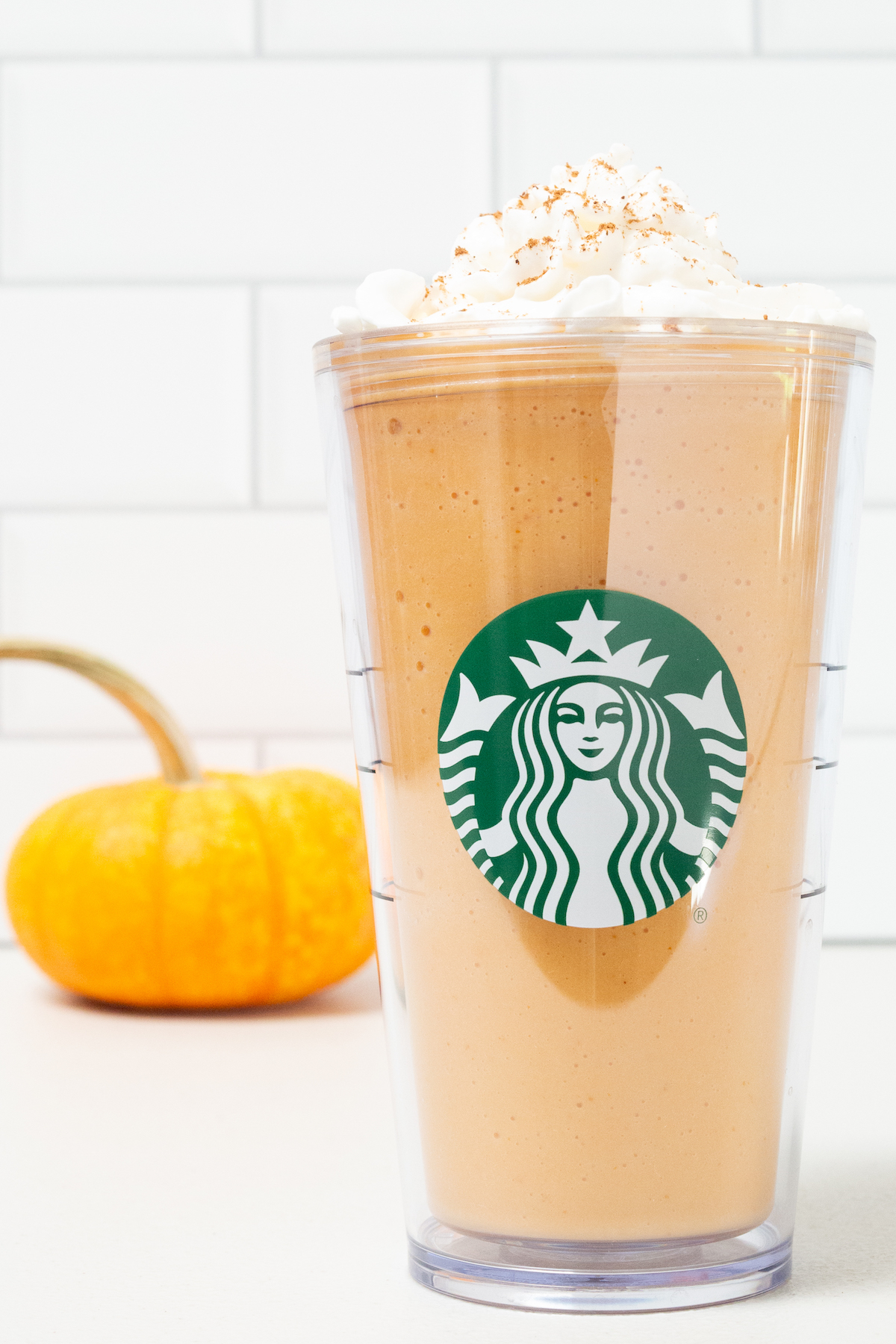 A tall plastic Starbucks tumbler is filled with pumpkin spice frappuccino that's been topped with whipped cream and pumpkin pie spice. A small orange pumpkin is out of focus in the background.