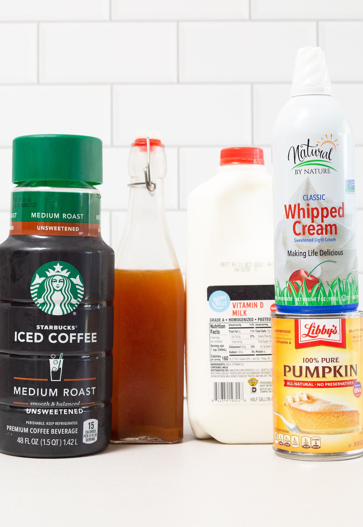 The ingredients to make a pumpkin spice frappuccino sit on a white background - iced coffee, pumpkin spice syrup, whole milk, canned pumpkin, and whipped cream