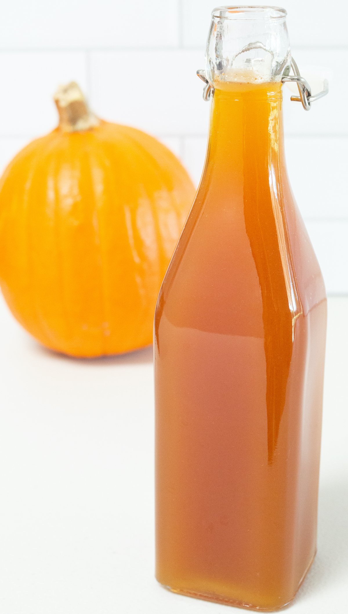 A brownish orange bottle of pumpkin spice syrup is in focus in the foreground, a pumpkin is out of focus in the background.