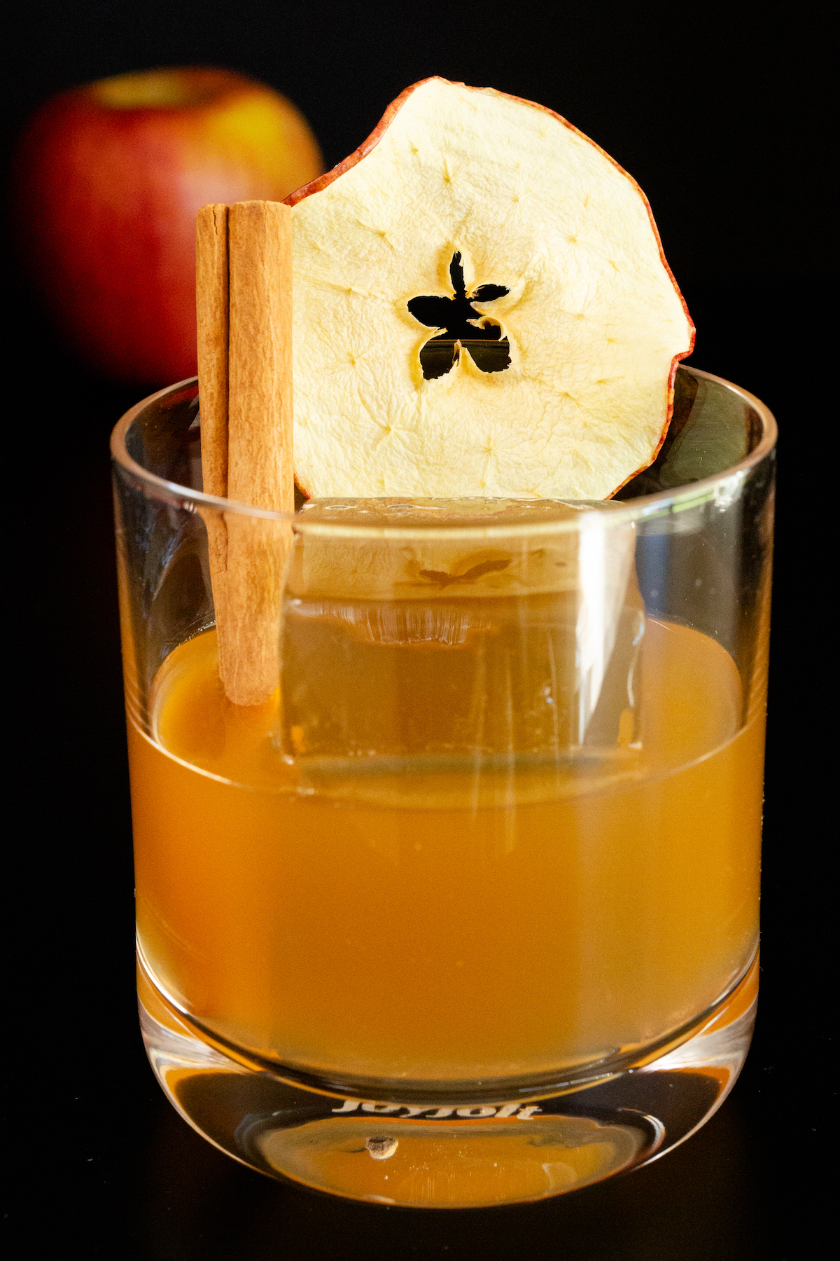 A lowball glass has been filled with a brown Apple Old Fashioned and a large clear block of ice. It is garnished with a dehydrated apple slice and a cinnamon stick.