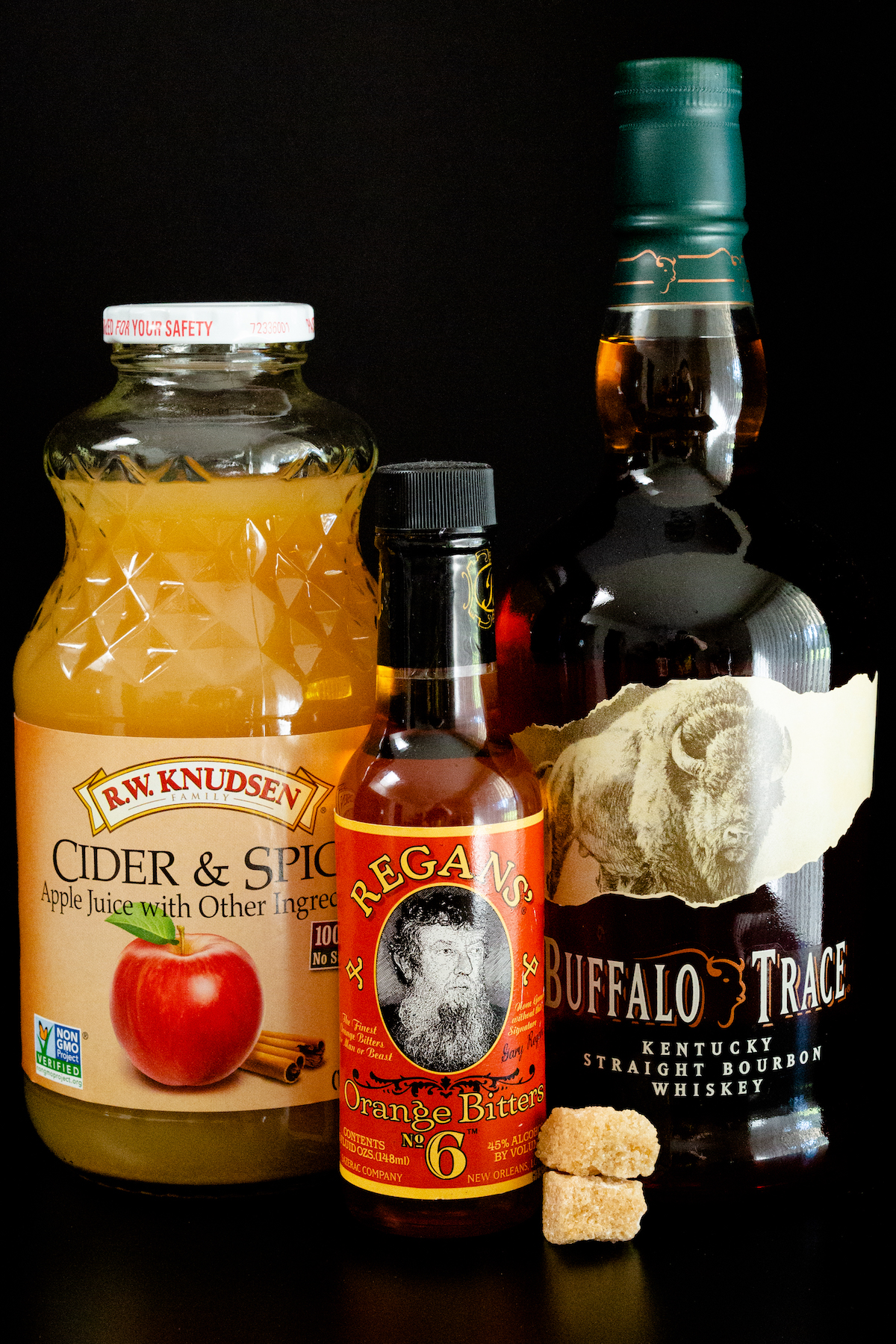 All the ingredients to make an apple old fashioned sit on a black background - a bottle of apple cider, bitter, bourbon, and two cubes of brown sugar.