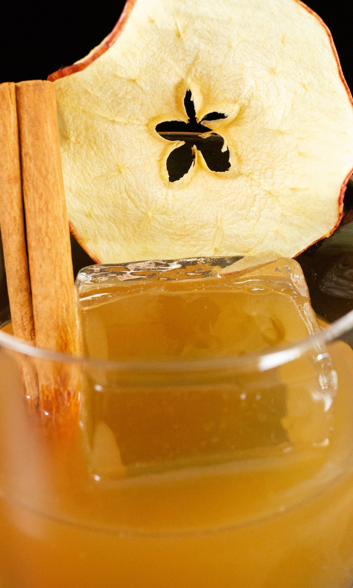 Close up of an apple old fashioned in a low ball glass. It is garnished by a whole dehydrated apple slice and cinnamon stick.