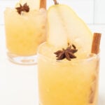 Two lowball glasses on a white background. They are filled with a yellow Thanksgiving cocktail that's garnished with a pear slice, star anise, and a cinnamon stick.