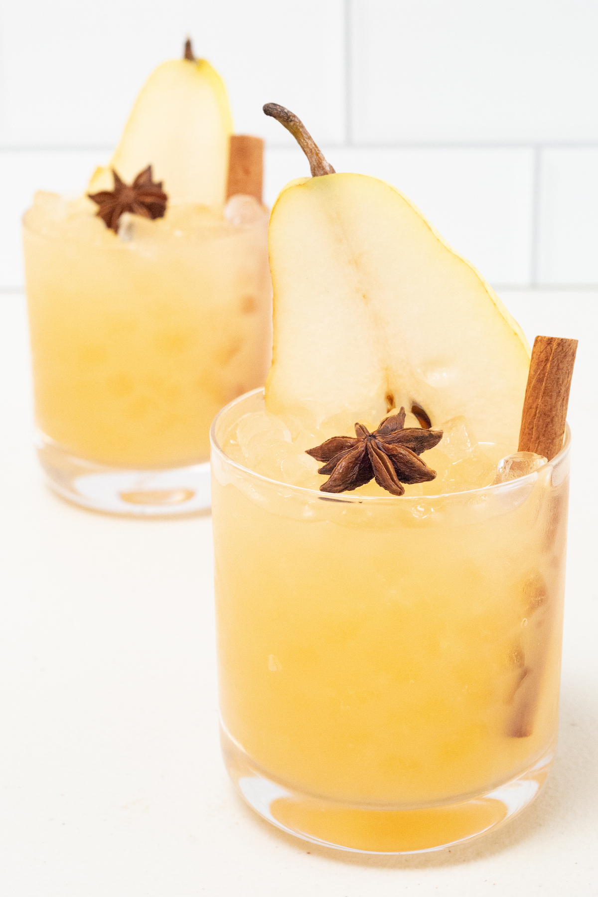 Two lowball glasses on a white background. They are filled with a yellow Thanksgiving cocktail that's garnished with a pear slice, star anise, and a cinnamon stick.
