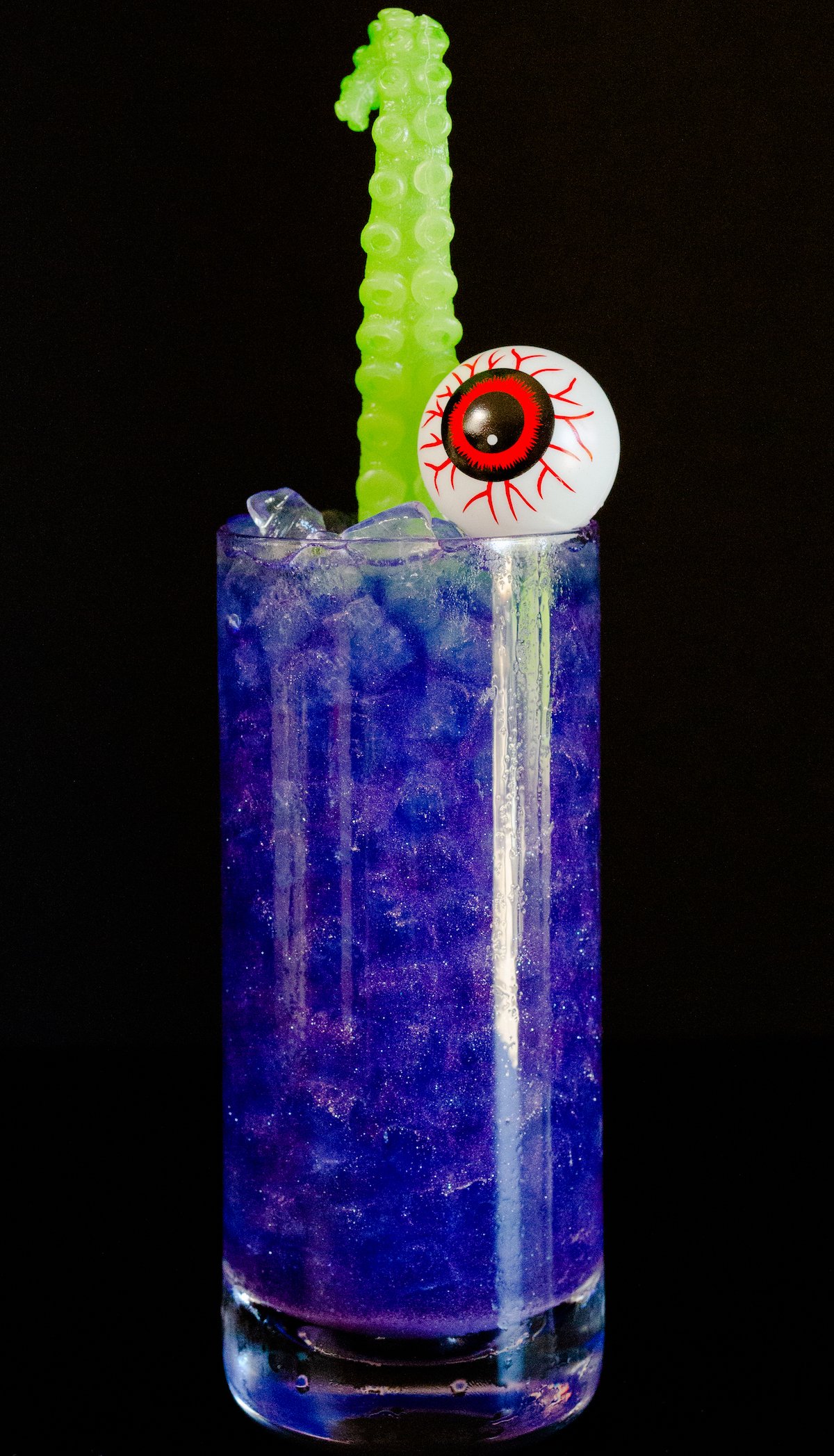 A highball glass filled with a bright shiny purple liquid sits on a black background. The drink is garnished with a green sea monster tentacle and plastic eyeball.