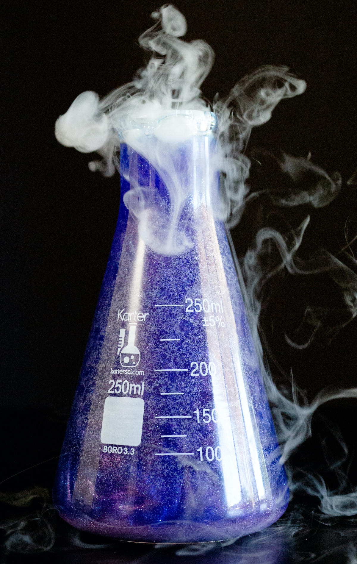 A scientific flask that's filled with a sparkling purple liquid sits on a black background. Smoke is rising out of the top.
